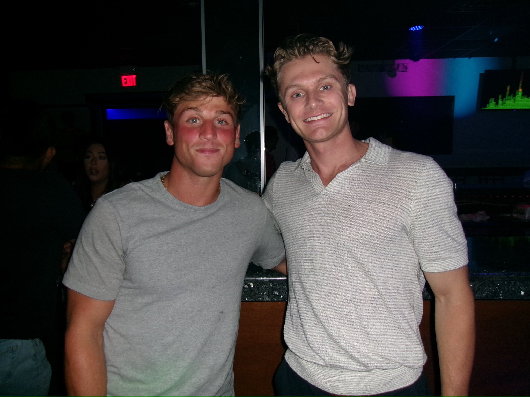 Two men posing together in a dark indoor setting with colorful lights.